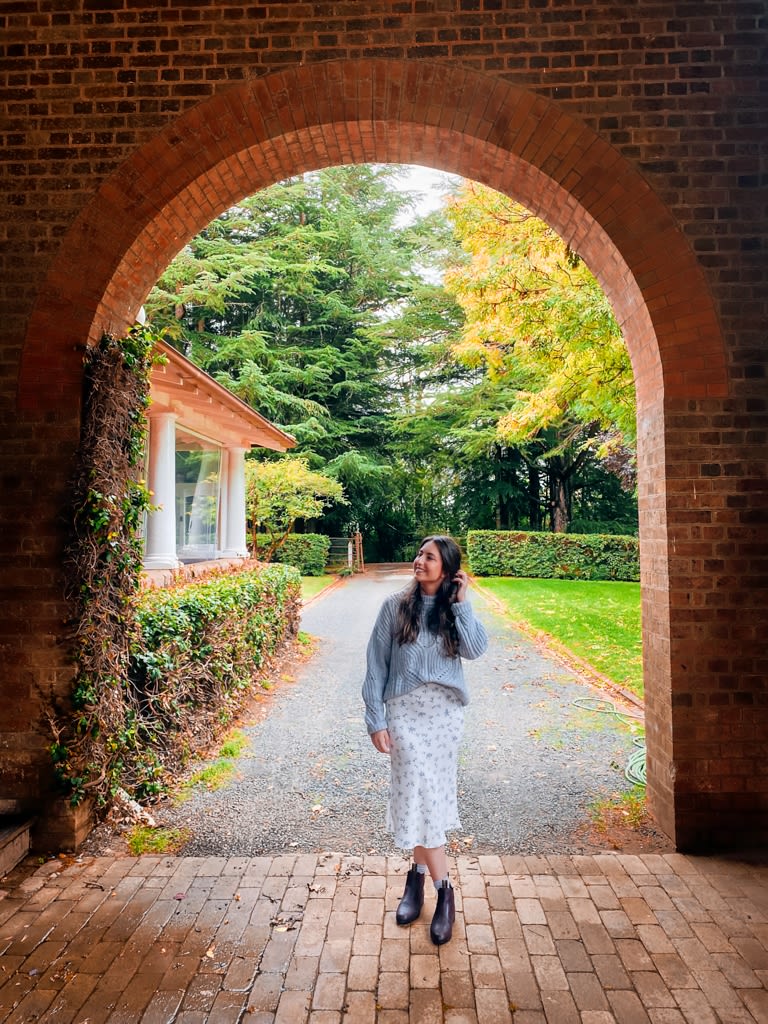Young woman in skirt and jumper stand in brick archway outside at a winery.  