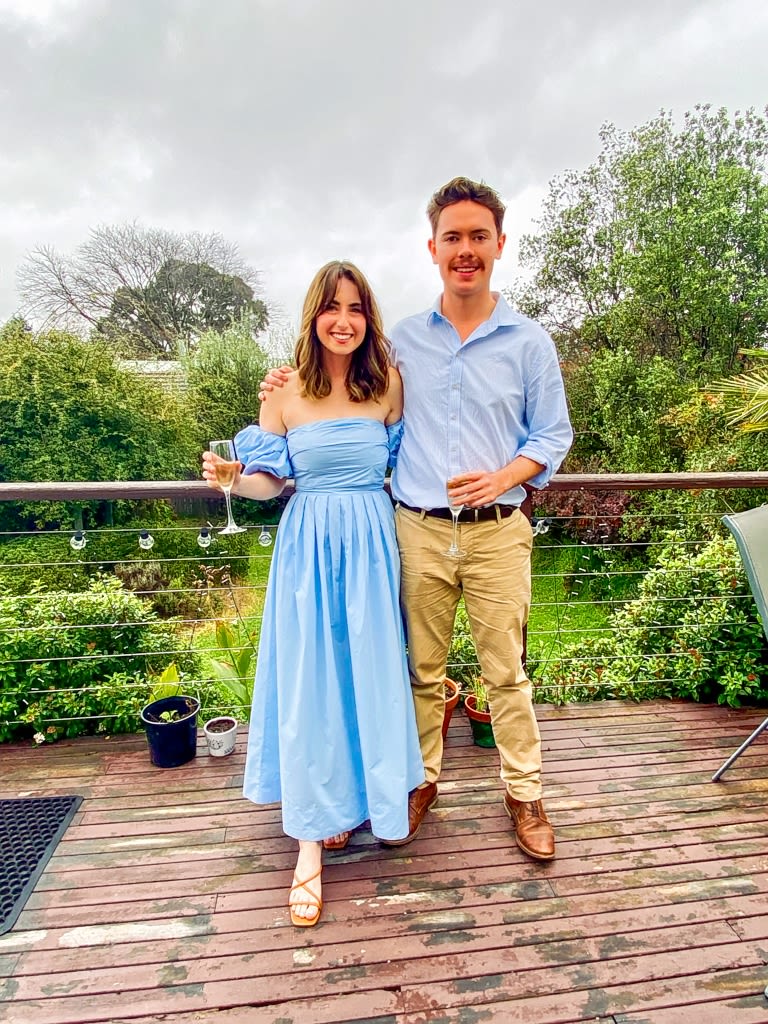 Young couple in formal wear pose for a photo holding wine glasses in a garden. 