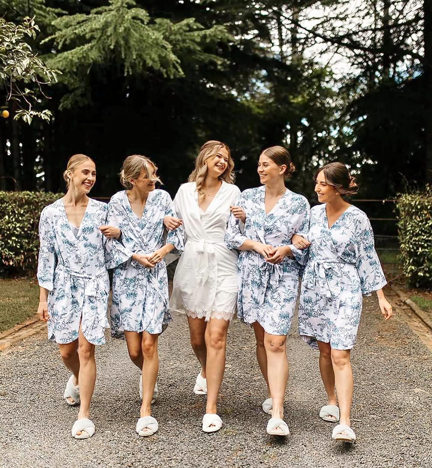 Bride and four bride maids walk along a path with arms interlocked wearing formal pyjamas. 