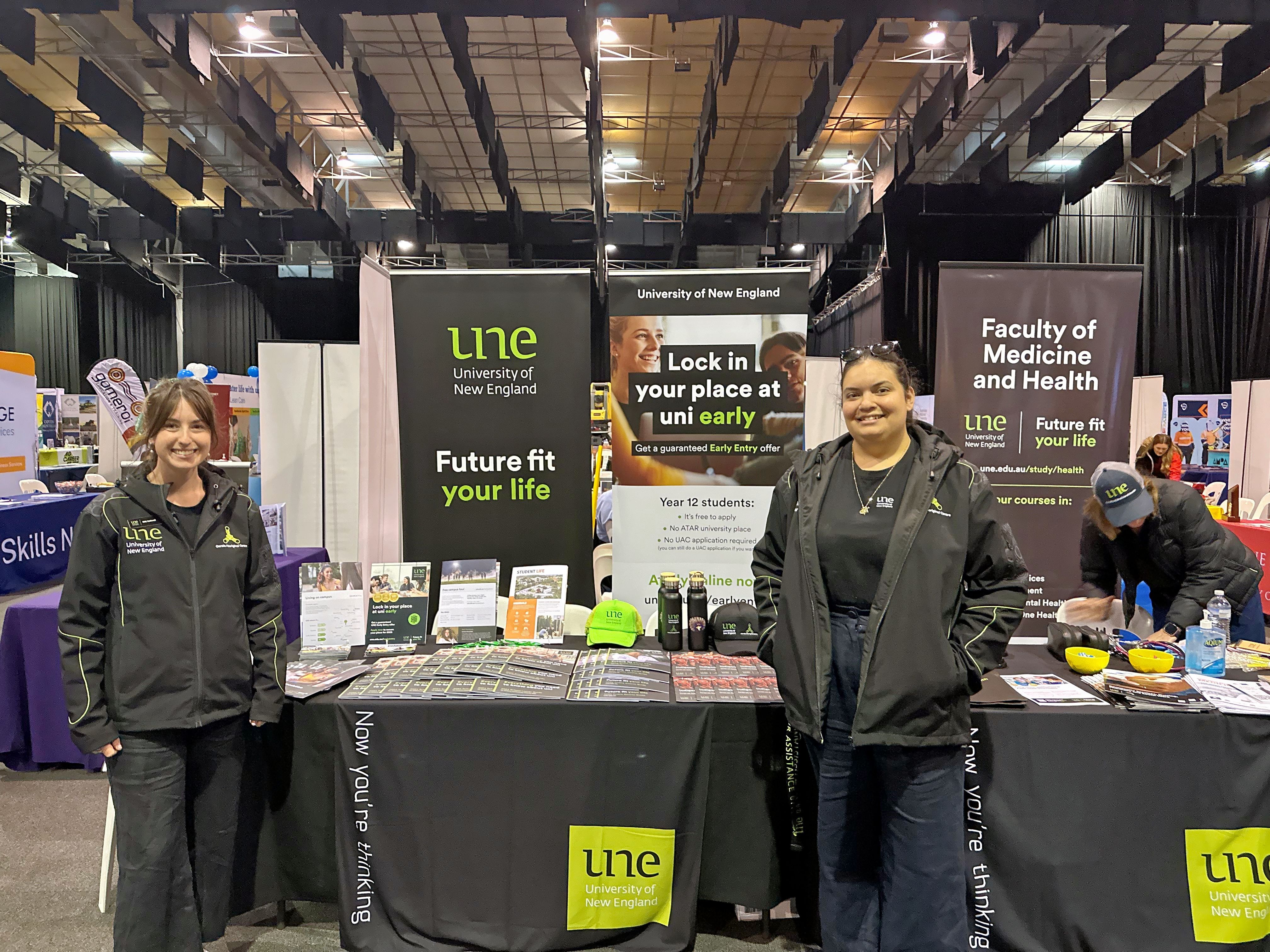 Two women in UNE uniforms pose for a photo in front of UNE stall at a careers expo. 