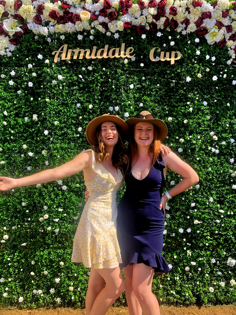 Two young women in dresses and hats pose for a photo at the races.
