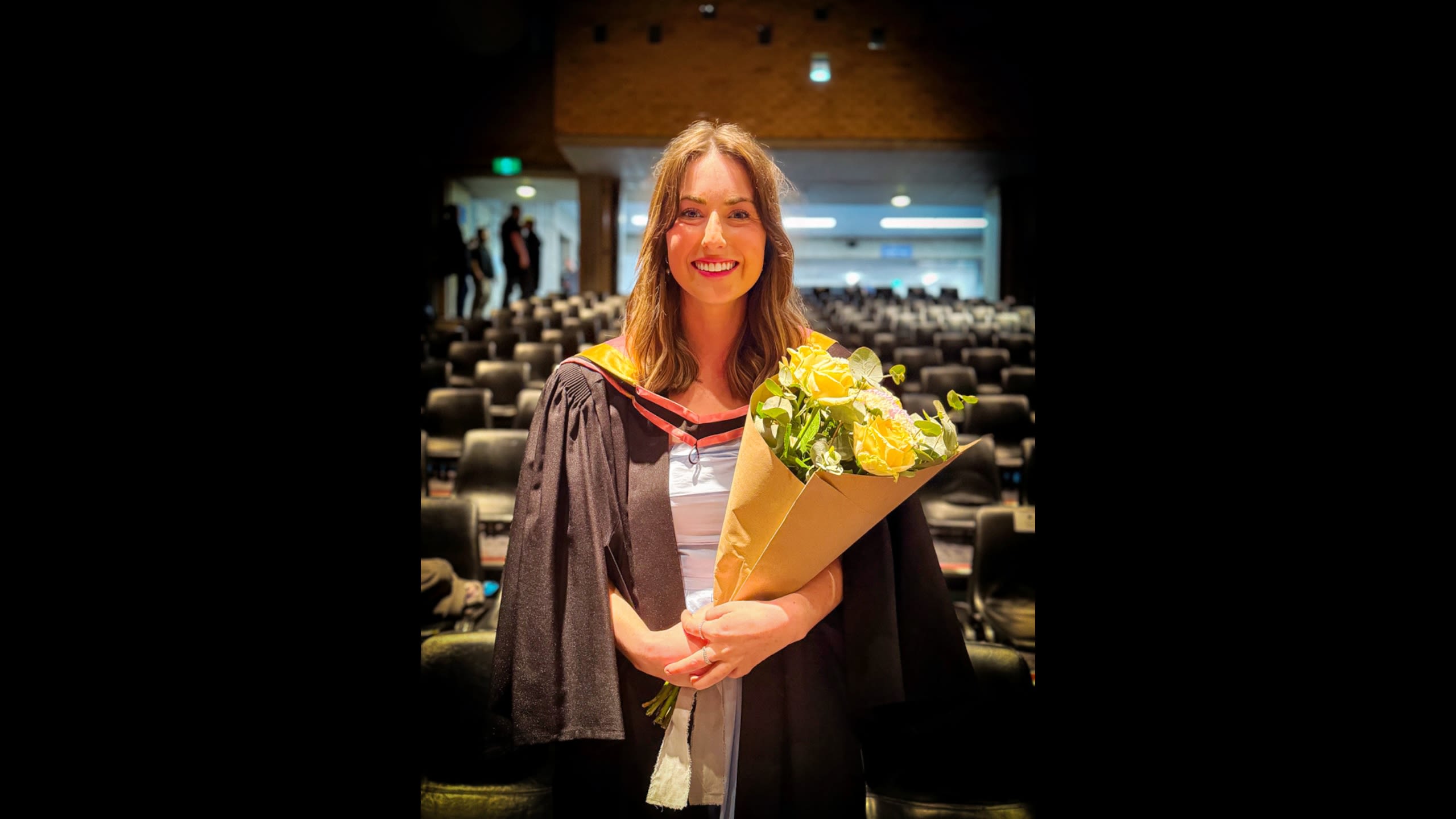 Young woman in graduation outfit inside an auditorium smiles for a photo holding a bouquet of flowers.