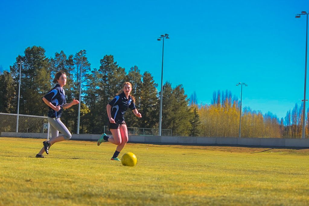 Two young women playing soccer chase after the ball on a soccer field.